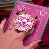 Heart E-reader and phone grip held by a hand with dark nail polish against a pink background