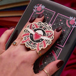 A detail shot of the black Sailor Swoon clingie on a kindle with a kindle sticker insert over a red fabric background. The kindle sticker insert design is a black, pink and red feminine bow design, the clingie featuring a flying heart and bows in the same colour palette.