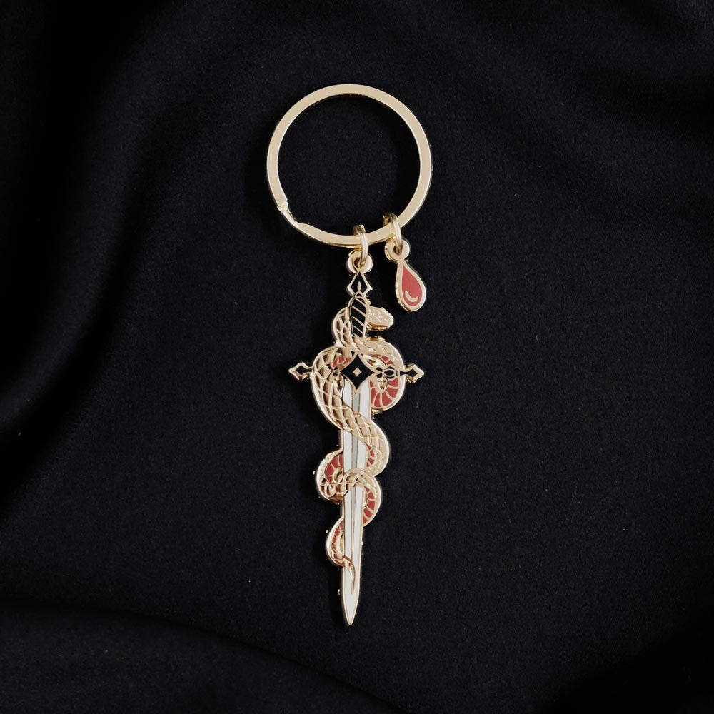 The romantasy inspired serpentine keyring by The Quirky Cup Collective sits on a black background. The gold keyring features black and red detailing and shows a snake curled around a sword alongside a drop of blood.