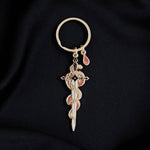 The romantasy inspired  serpentine keyring by The Quirky Cup Collective sits on a black background. The gold keyring features black and red detailing and shows a snake curled around a sword alongside a drop of blood.