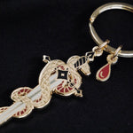 A detail shot of the Serpentine keyring by The Quirky Cup Collective sits on a black background. The gold keyring features black and red detailing and shows a snake curled around a sword alongside a drop of blood.