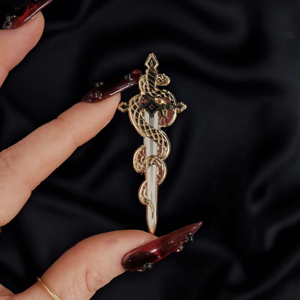 The fantasy book inspired Serpentine enamel pin by The Quirky Cup Collective sits on a black background, red nailed fingers reaching towards it. The gold pin features black and red detailing and shows a snake curled around a sword alongside a drop of blood.