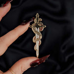 The fantasy book inspired Serpentine enamel pin by The Quirky Cup Collective sits on a black background, red nailed fingers reaching towards it. The gold pin features black and red detailing and shows a snake curled around a sword alongside a drop of blood.