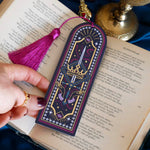 The blue Oathbreaker bookmark is an arch shaped bookmark with a pink tassle. The blue, pink and white design features hand, crown, sword and snake motifs and gold foiling for a luxe romantasy aesthetic edge. the bookmark sits in an open book on a shadowy blue fabric background.
