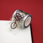 Swords and Serpents transparent sticker washi tape by The Quirky Cup Collective sits on a journal on a red table. The fantasy book inspired washi tape is red, black, white and fold and functions like a set of individual stickers featuring motifs of snakes, swords, crowns, dragons and more.