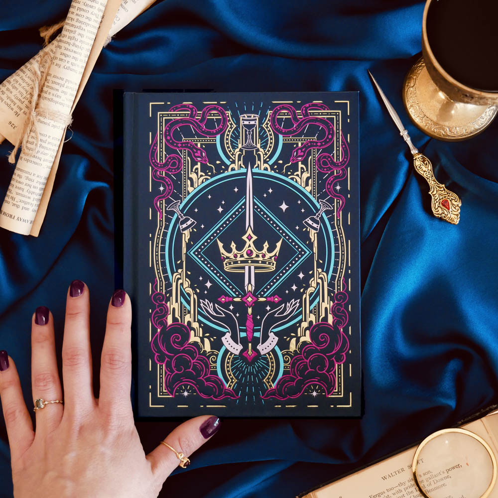 A close up shot of the cover of the blue 'Oath Breaker' journal by The Quirky Cup Collective. The deep blue cover shows the pink, teal and gold foiling and motifs of snakes, swords, goblets and crowns for a fantasy book aesthetic. The book is on a blue fabric background and is surrounded by a gold goblet and scrolls.