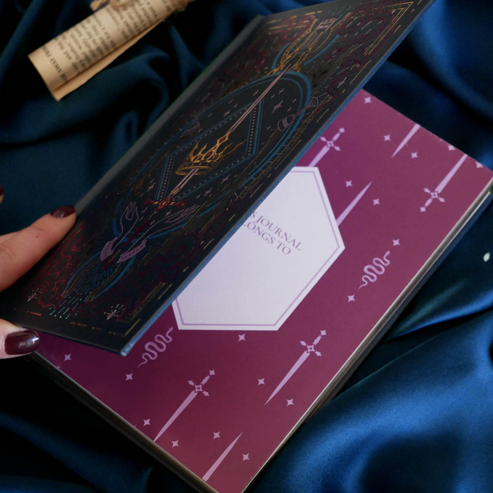 In a dark scene, the front page of a blue Quirky Cup Collective reading journal is held open to show a nameplate on a lush pink spread with snake and sword motifs. The journal is help open by a hand with long red nails on a deep blue fabric background surrounded by scrolls.