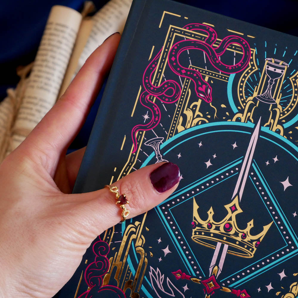 A close up shot of the cover of the blue 'Oath Breaker' journal by The Quirky Cup Collective. The deep blue cover shows the pink, teal and gold foiling and motifs of snakes, swords, goblets and crowns for a fantasy book aesthetic.