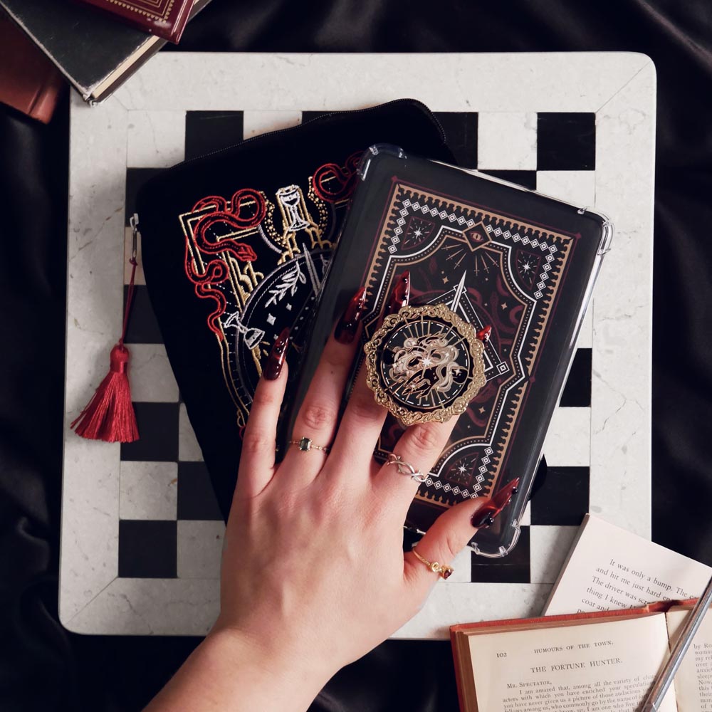 The Serpents and Swords e-reader bundle shows a e-reader sleeve and a kindle that has a kindle sticker insert and the magic weaver clingie kindle grip. A hand holds the kindle in front of a chess board on a black table covered in books. The romantasy inspired products are all black with red, white and gold detailing and all feature motifs of a swords, crowns, snakes and hands with swiling magic.