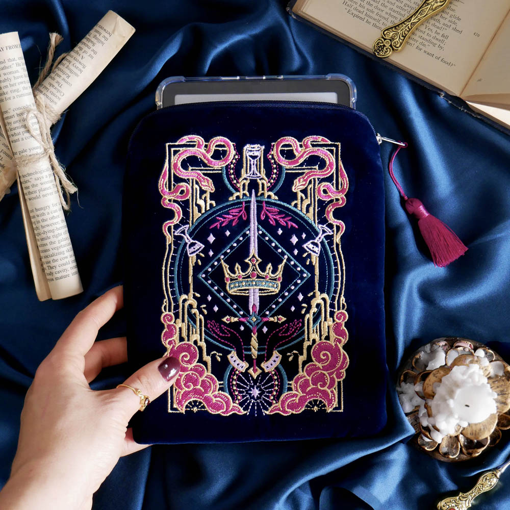 The Blue Serpents & Swords E-Reader Sleeve. On a blue fabric background, a hand holds the embroidered kindle case with an e-reader sticking out the top. The case features a pink tassel and pink, gold and blue toned embroidery showing a sword, crown and snake motif.