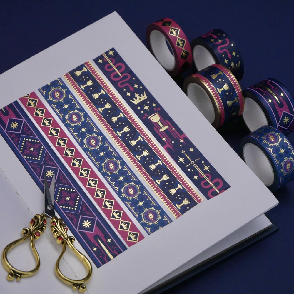 The full set of Serpents and Swords washi tapes, a romantasy book inspired collection by The Quirky Cup Collective, are show on a white journal on a blue table. The five washi tapes are in shades of pink and blue with intricate gold foiling. The designs feature motifs of snakes, crowns, swords, goblets , stars and serpents and perfectly match the fantasy book aesthetic.