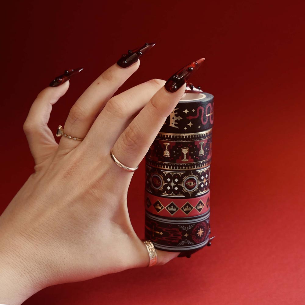 The full set of Serpents and Swords washi tapes, a fantasy book inspired collection by The Quirky Cup Collective, are held in a hand with intricate red nails in front of a red backround. The five washi tapes are in shades of red, black and white with intricate gold foiling. The designs feature motifs of snakes, crowns, swords, goblets , stars and serpents and perfectly match the fantasy book aesthetic.