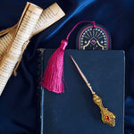 The blue Oathbreaker bookmark is an arch shaped bookmark with a pink tassle. The blue, pink and white design features hand, crown, sword and snake motifs and gold foiling for a luxe romantasy aesthetic edge. The bookmark is peaking our from a navy coloured book on a blue fabric background surrounded by scrolls and a gold letter opener.