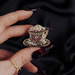 black red and gold teacup with snake detail enamel pin held by a hand against a dark background