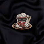 black red and gold teacup with snake detail enamel pin held by a hand against a dark background