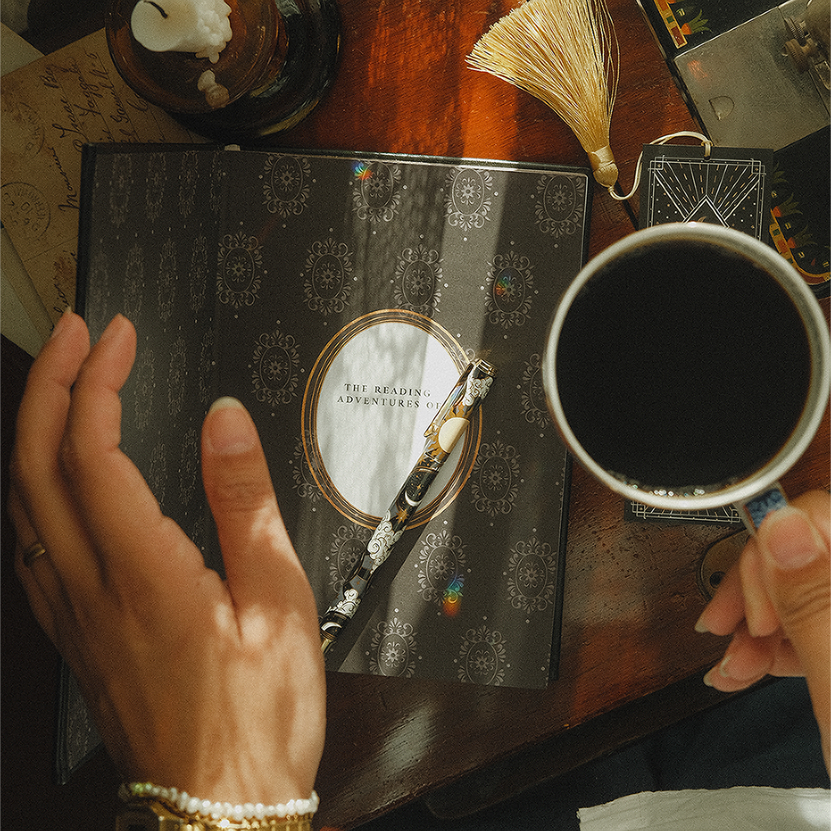 Ambassador image of an open Reading Journal page, hands holding a cup of tea next to a Quirky Cup Collective pen and bookmark.