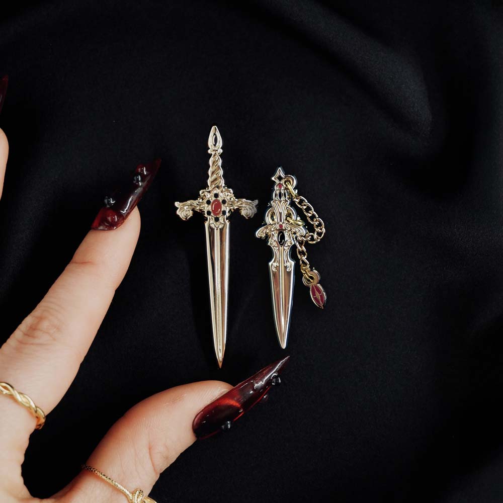 Gold red and black dagger and sword enamel pin held by a hand against a dark background