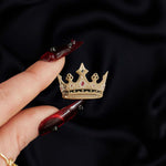 Gold black and red crown enamel pin held by a hand against a dark background