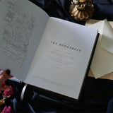 A hand with dark red nail polish and a delicate ring rests on the cover of a Black Reading Journal by The Quirky Cup Collective, showcasing its ornate gold and white embossed design and the words 'Reading Journal' in the center. The journal lies on a smooth black satin fabric, with dried red roses and a decorative candle holder nearby, creating a luxurious and elegant ambiance