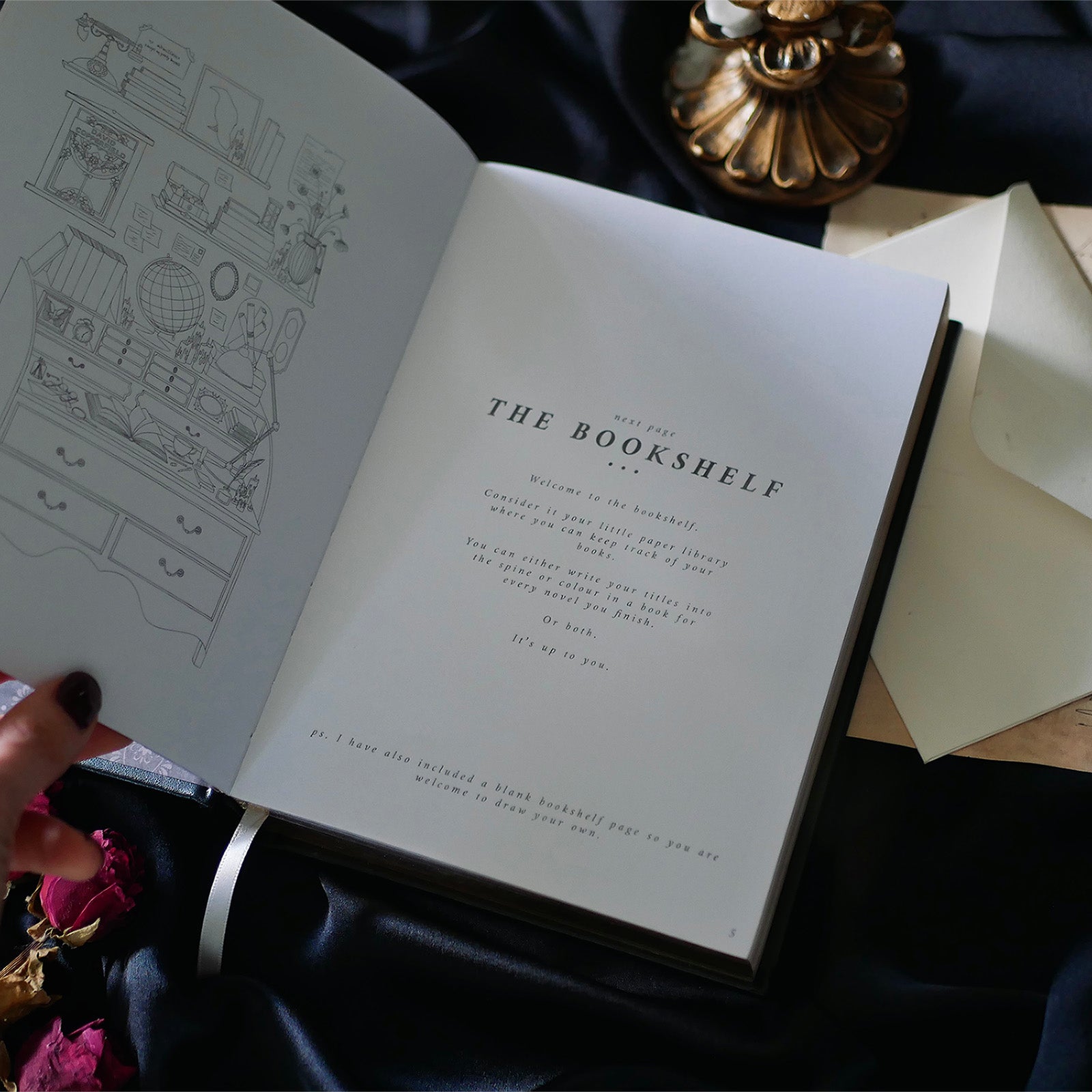 A hand with dark red nail polish and a delicate ring rests on the cover of a Black Reading Journal by The Quirky Cup Collective, showcasing its ornate gold and white embossed design and the words 'Reading Journal' in the center. The journal lies on a smooth black satin fabric, with dried red roses and a decorative candle holder nearby, creating a luxurious and elegant ambiance