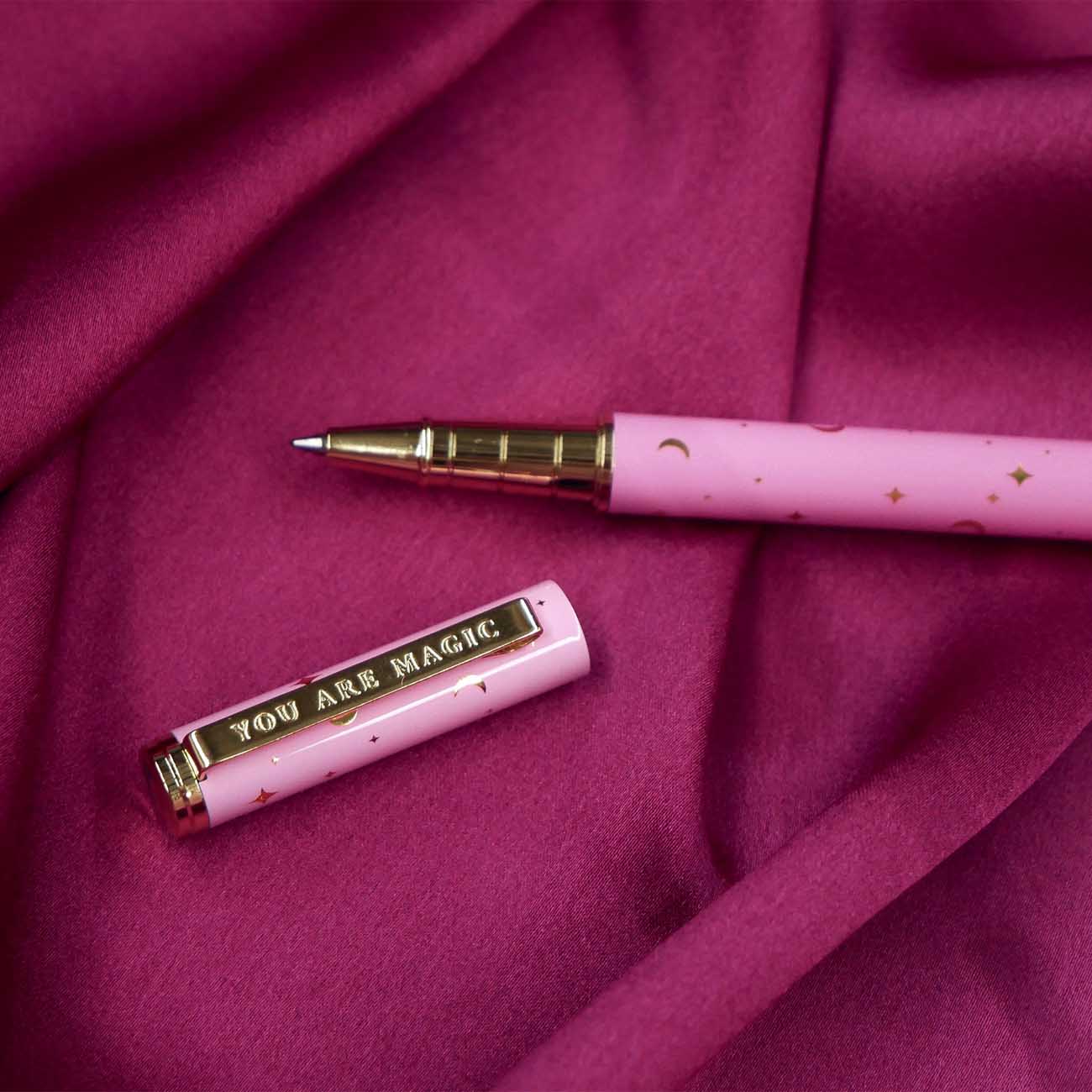 A close up of the You Are Magic pen in bubblegum pink. On a pink fabric background, the pink pen features stars and moons in gold and the phrase 'You are magic' on the gold pen clip.