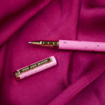 A close up of the You Are Magic pen in bubblegum pink. On a pink fabric background, the pink pen features stars and moons in gold and the phrase 'You are magic' on the gold pen clip.