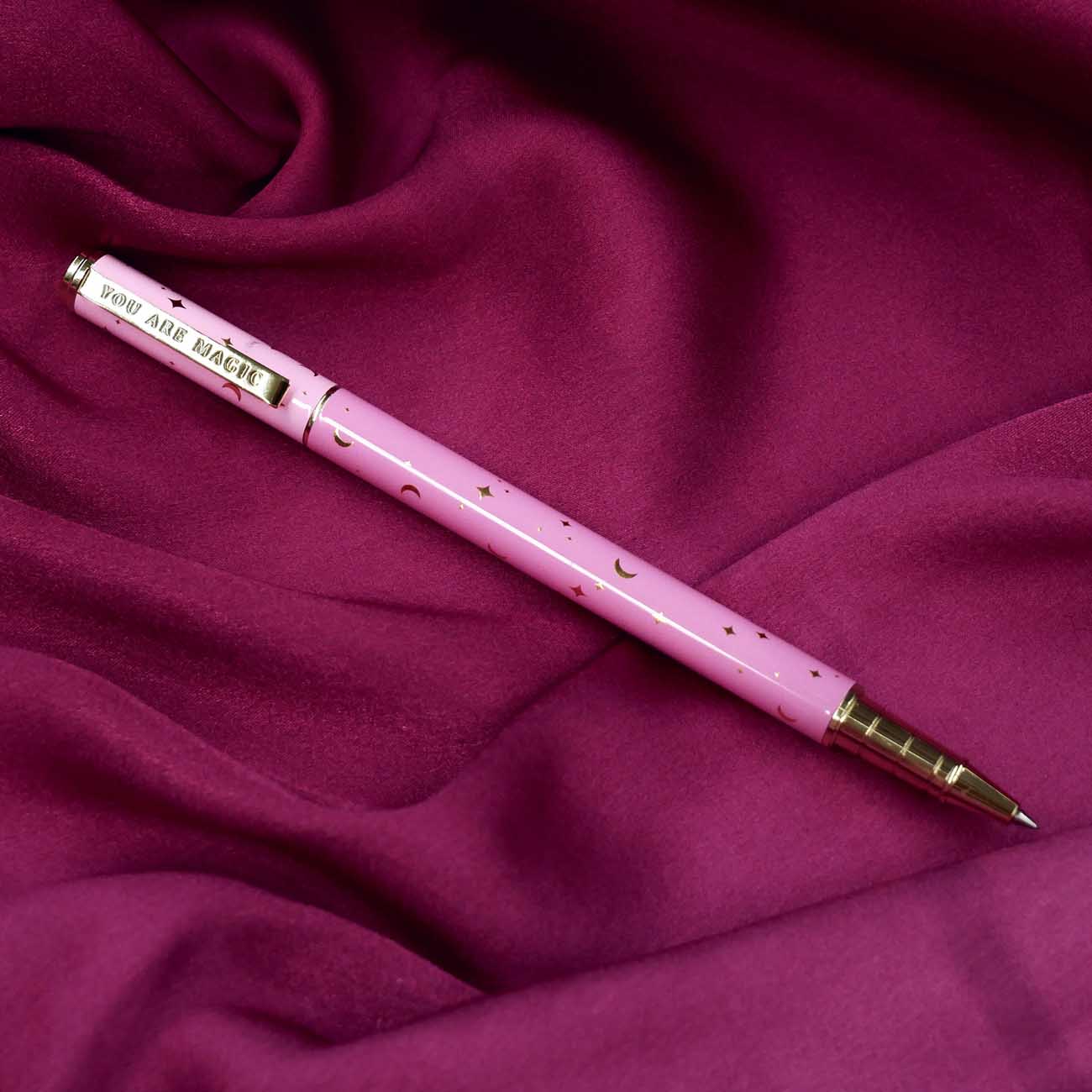 A close up of the You Are Magic pen in bubblegum pink. On a pink fabric background, the pink pen features stars and moons in gold and the phrase 'You are magic' on the gold pen clip.