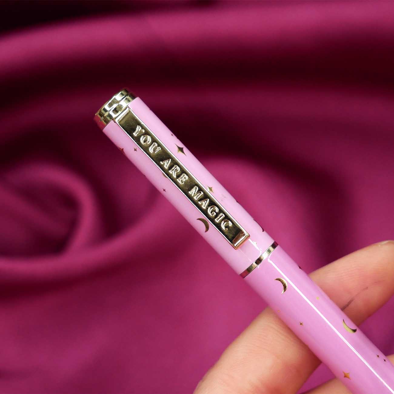 A close up of the You Are Magic pen in bubblegum pink. On a pink fabric background, the pink pen features stars and moons in gold and the phrase 'You are magic' on the gold pen clip.