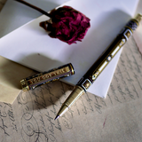 A hand holding the Black You Got This Pen by The Quirky Cup Collective, showcasing its intricate gold and white floral and geometric barrel design on a black background. The pen is positioned on top of a group of letters and envelopes with dried red roses creating a moody, vintage-inspired scene.
