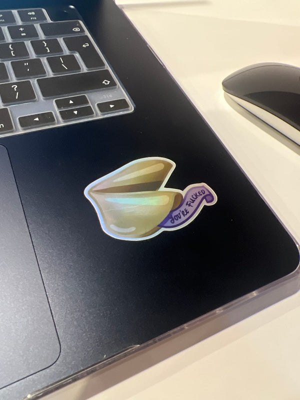 One of the The Quirky Cup Collection vinyl stickers in use. On the edge of a laptop is a small holographic vinyl sticker of a fortune cookie with the message 'you're fucked'