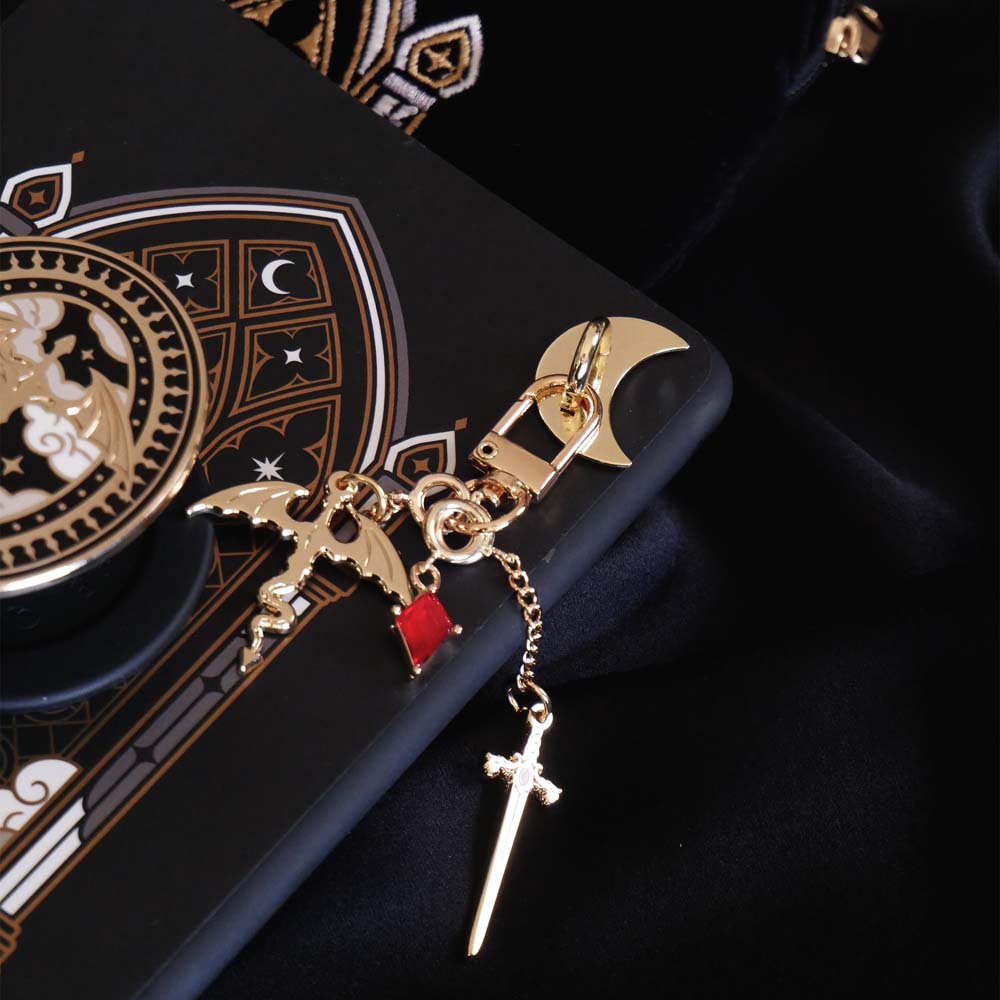 The detail of the gold Dragon rider Charmlet (A mix and match accessory for decorating your kindle) by The Quirky Cup Collective attached to a kindle with the matching kindle sticker insert design sits on a black fabric background. The design featured a gold clasp and multiple charms hanging from different length detachable chains including a dragon, a sword and a red gem. The charmlet is attached to the device with a gold moon charm anchor.