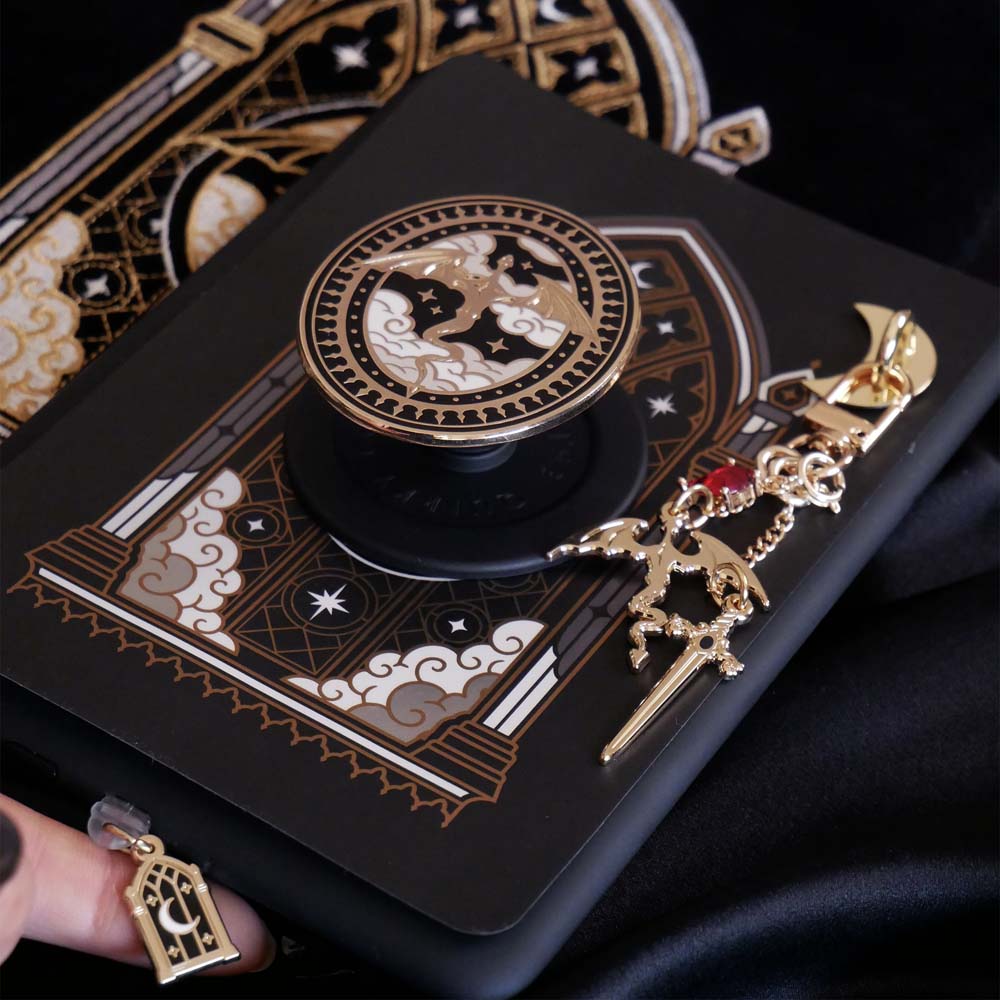 The Dragon Lore E-Reader decor bundle by The Quirky Cup Collective sits on a black fabric background surrounded by books, a hand with ornate pastel nails holding it. The black and gold set includes an embroidered e-reader case, kindle insert sticker, Clingie (imagine an enamel pop socket or decorative phone grip), Charmlet (An accessory with multiple charms attached) and a dust plug. All of the elements feature a design of a gold dragon flying through clouds in front of an arched stained glass window motif.
