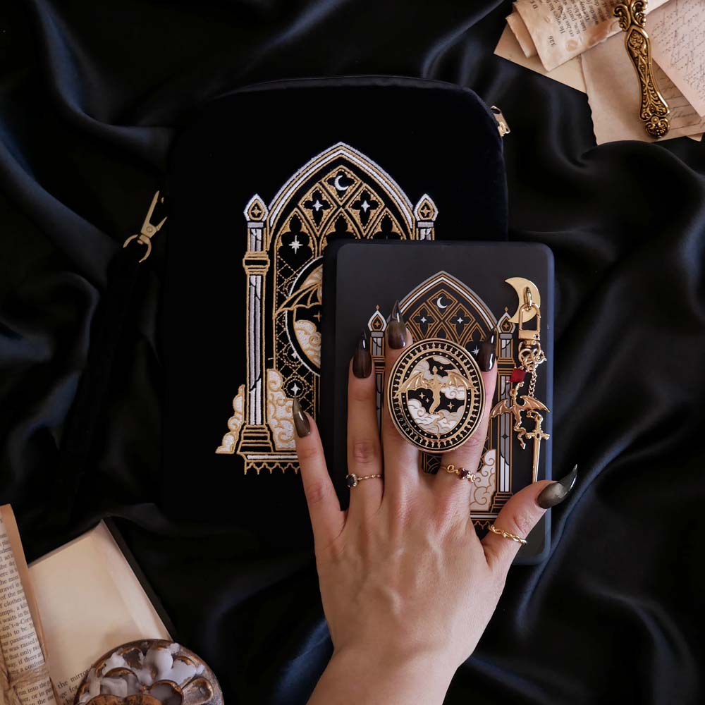 The Dragon Lore E-Reader decor bundle by The Quirky Cup Collective sits on a black fabric background surrounded by books, a hand with ornate pastel nails holding it. The black and gold set includes an embroidered e-reader case, kindle insert sticker, Clingie (imagine an enamel pop socket or decorative phone grip), Charmlet (An accessory with multiple charms attached) and a dust plug. All of the elements feature a design of a gold dragon flying through clouds in front of an arched stained glass window motif.