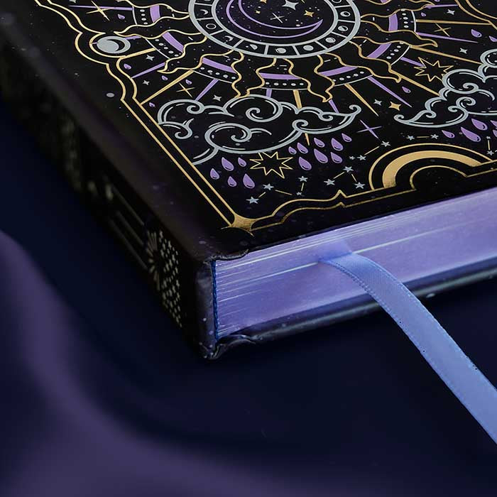 Journal with celestial cover sits on a purple silky background showing the purple foiled edge of the journal and a ribbon bookmark sticking our of the bottom pages