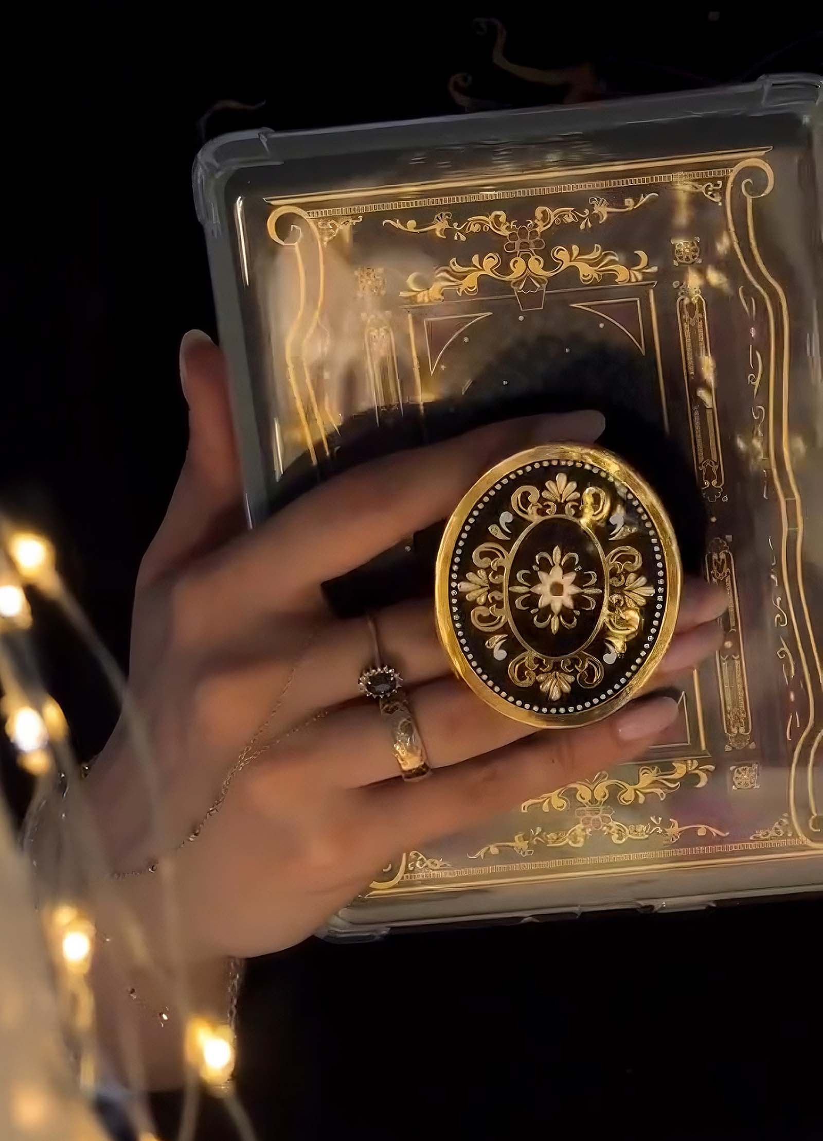 Decorative gold and black phone grip held by a hand with a ring, against a dark background.