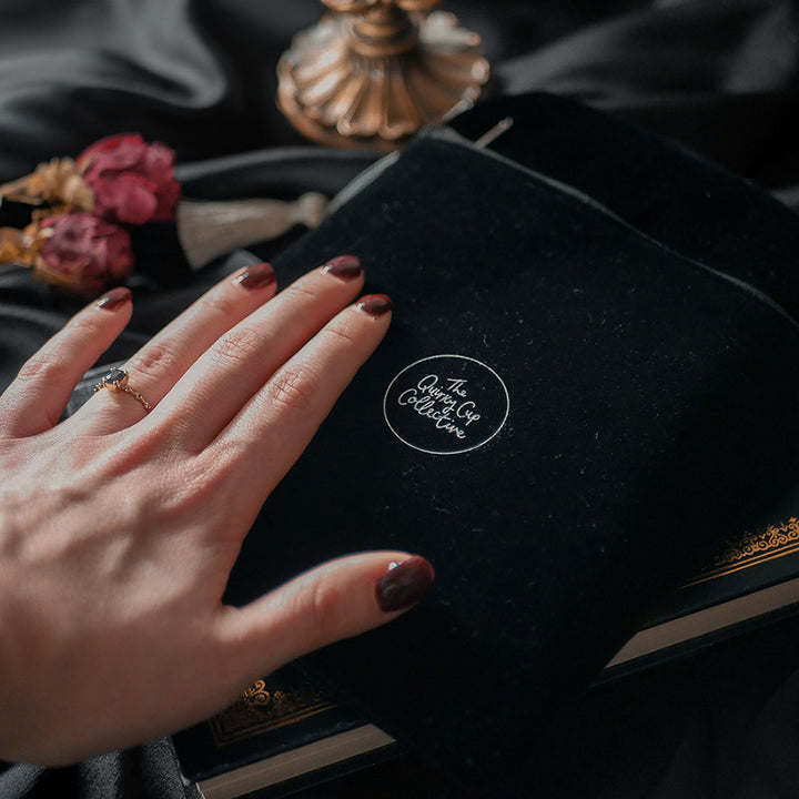 A hand holding a luxurious black velvet book sleeve with an intricate gold and silver embroidered design by The Quirky Cup Collective. The Kindle & E-reader sleeve, featuring a decorative tassel zipper, holds an e-reader, peeking out from the top. The background is a smooth black satin fabric, with dried roses and rolled-up book pages placed beside the  Kindle & E-reader sleeve, adding a vintage and elegant aesthetic to the scene.
