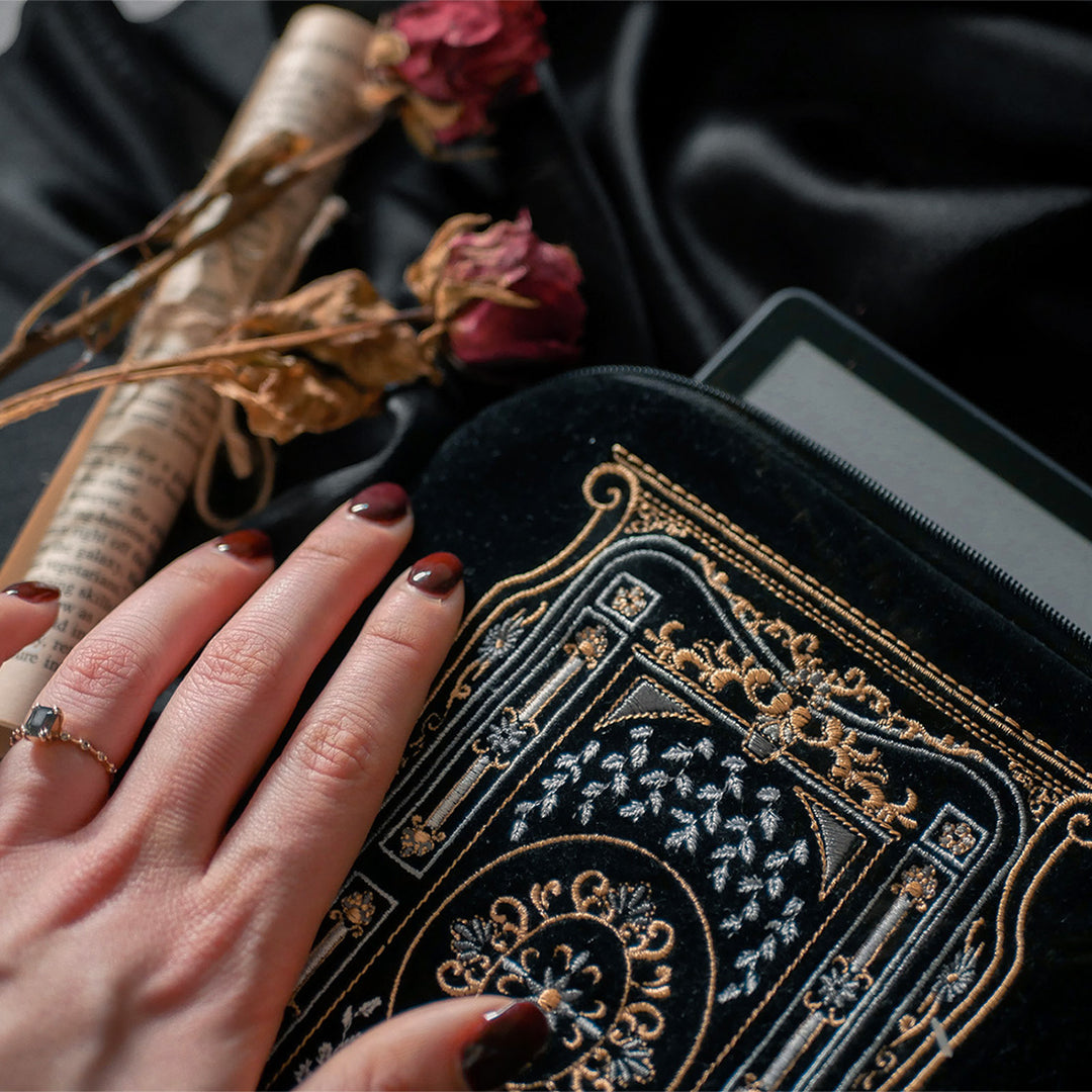 A hand holding a luxurious black velvet book sleeve with an intricate gold and silver embroidered design by The Quirky Cup Collective. The Kindle & E-reader sleeve, featuring a decorative tassel zipper, holds an e-reader, peeking out from the top. The background is a smooth black satin fabric, with dried roses and rolled-up book pages placed beside the  Kindle & E-reader sleeve, adding a vintage and elegant aesthetic to the scene.