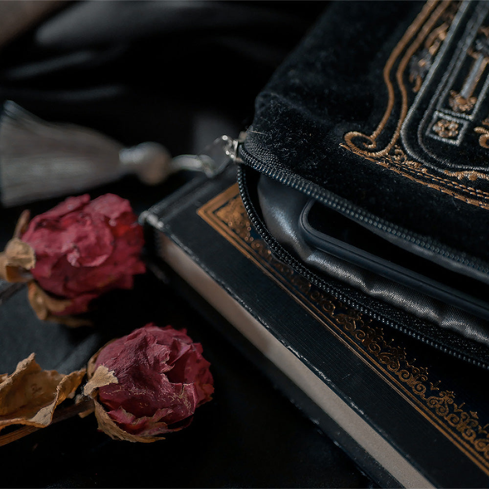 A close-up view of a luxurious black velvet sleeve with intricate gold embroidery, partially unzipped to reveal the edge of an e-reader. The sleeve features a decorative tassel zipper and is placed on top of a black book with a gold-embossed cover. Two dried red roses are elegantly positioned next to the sleeve, creating a vintage and refined aesthetic against the dark, soft background.