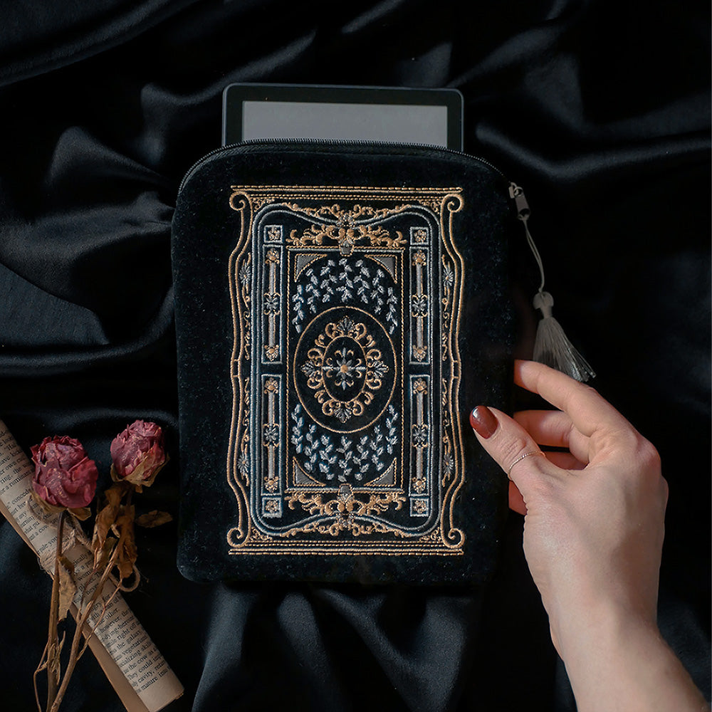 A hand holding a luxurious black velvet book sleeve with an intricate gold and silver embroidered design by The Quirky Cup Collective. The sleeve, featuring a decorative tassel zipper, holds an e-reader, peeking out from the top. The background is a smooth black satin fabric, with dried roses and rolled-up book pages placed beside the sleeve, adding a vintage and elegant aesthetic to the scene.