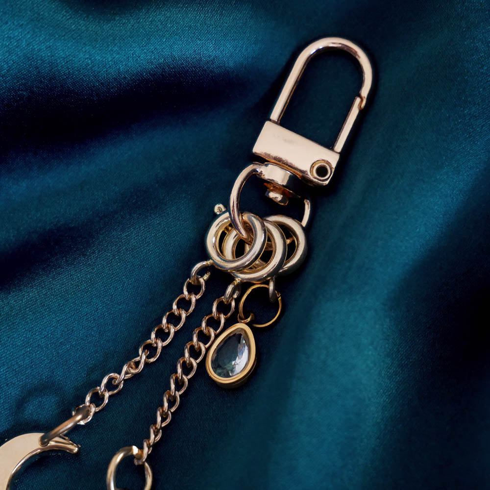 A detail of the gold Luna Moth Charmlet (A mix and match accessory for customising your kindle in a romantasy aesthetic) by The Quirky Cup Collective sits on a dark green fabric background. The design featured a gold clasp and multiple charms hanging from different length detachable chains including a luna moth in multiple shades of green attached to a star, a moon and a jade coloured gem.