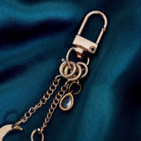 A detail of the gold Luna Moth Charmlet (A mix and match accessory for customising your kindle in a romantasy aesthetic) by The Quirky Cup Collective sits on a dark green fabric background. The design featured a gold clasp and multiple charms hanging from different length detachable chains including a luna moth in multiple shades of green attached to a star, a moon and a jade coloured gem.