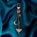 The gold Luna Moth Charmlet (A mix and match accessory for customising your kindle in a romantasy aesthetic) by The Quirky Cup Collective sits on a dark green fabric background. The design featured a gold clasp and multiple charms hanging from different length detachable chains including a luna moth in multiple shades of green attached to a star, a moon and a jade coloured gem.