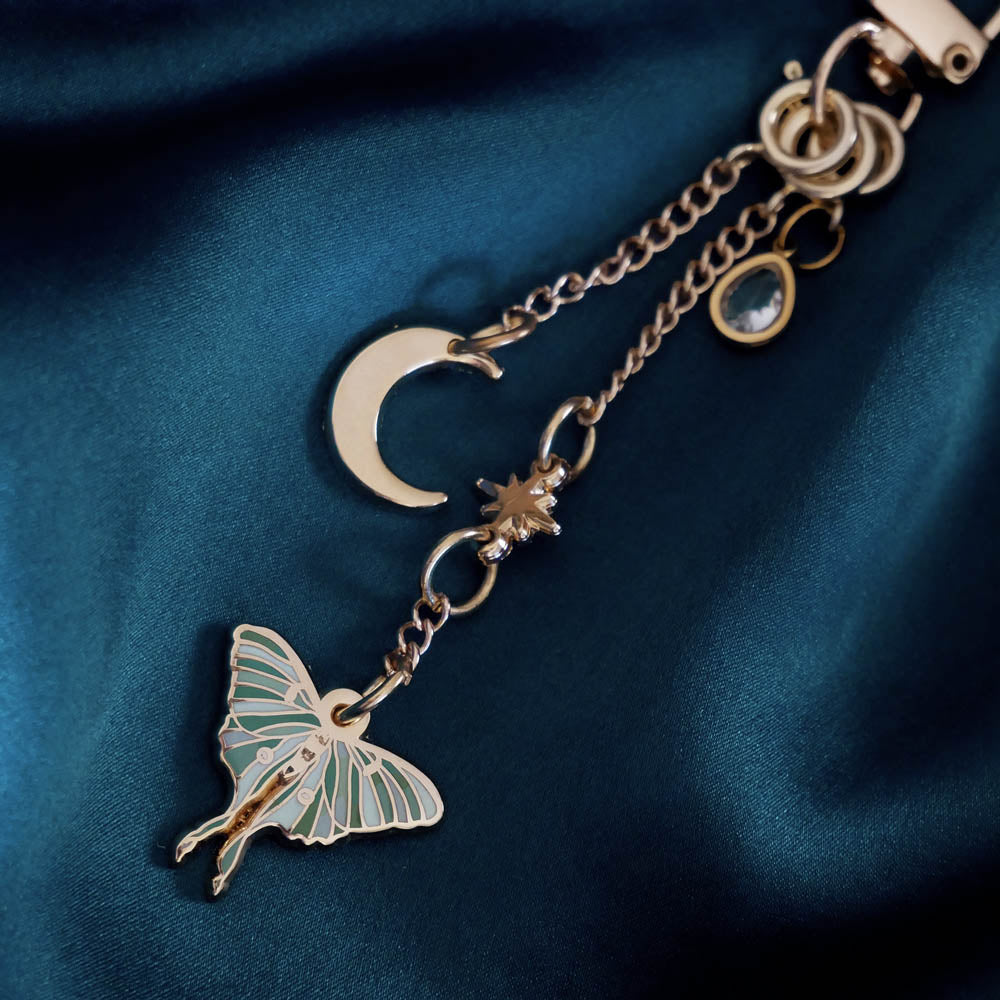 A detail of the gold Luna Moth Charmlet (A mix and match accessory for customising your kindle in a romantasy aesthetic) by The Quirky Cup Collective sits on a dark green fabric background. The design featured a gold clasp and multiple charms hanging from different length detachable chains including a luna moth in multiple shades of green attached to a star, a moon and a jade coloured gem.