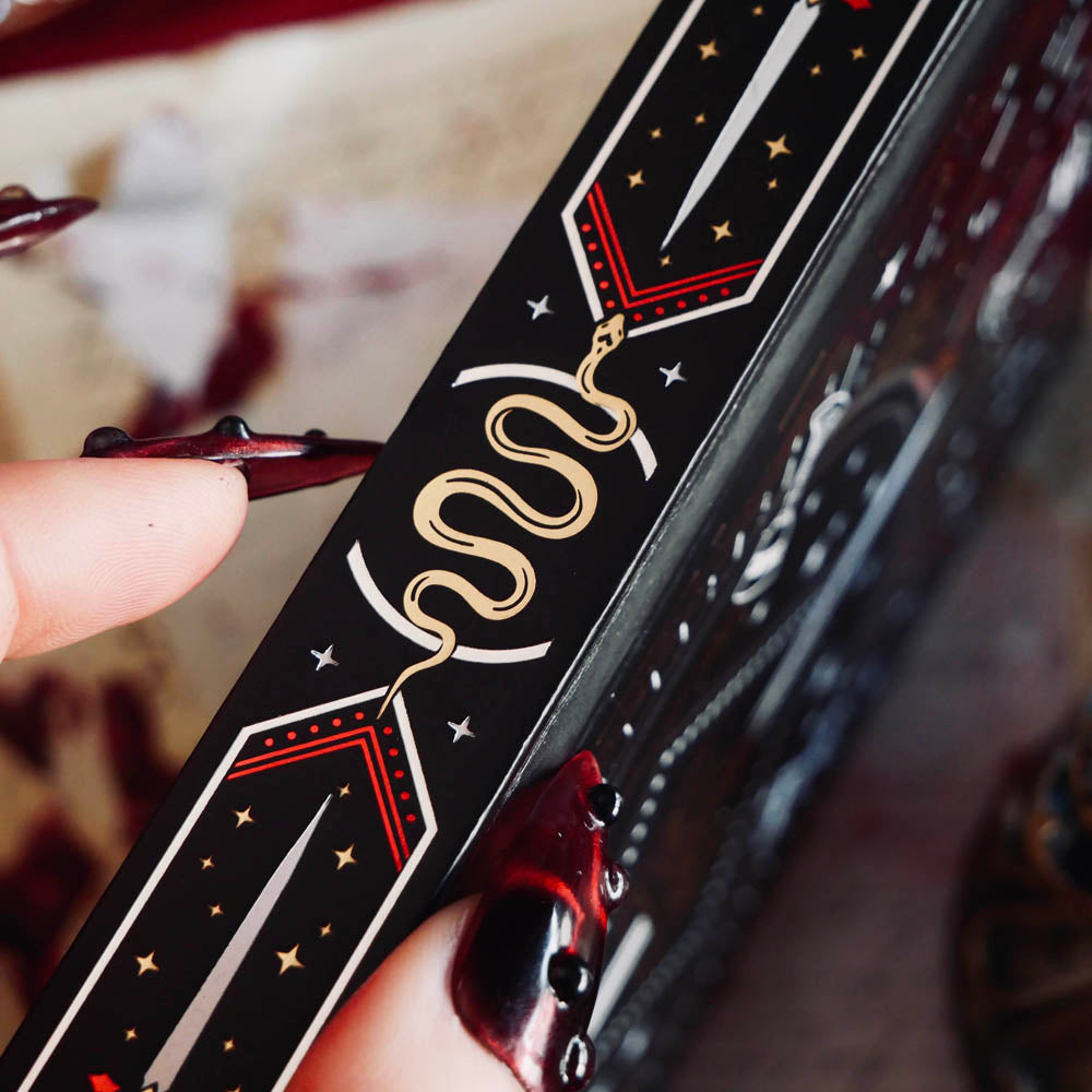 A detail of the spine of the Oath Breaker Journal sits on a red fabric background surrounded by handwritten notes, a candle and a crown. The black journal features intricate gold, red and white foiling with motifs inspired by fantasy books such as crowns, swords, goblets and snakes.