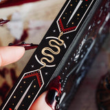 A detail of the spine of the Oath Breaker Journal sits on a red fabric background surrounded by handwritten notes, a candle and a crown. The black journal features intricate gold, red and white foiling with motifs inspired by fantasy books such as crowns, swords, goblets and snakes.