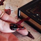 A detail of the Oath Breaker Journal sits surrounded by handwritten notes, a candle and a crown. The black journal features intricate gold, red and white foiling with motifs inspired by fantasy books such as crowns, swords, goblets and snakes. The gilded gold pages are shown with the red placeholder ribbon.