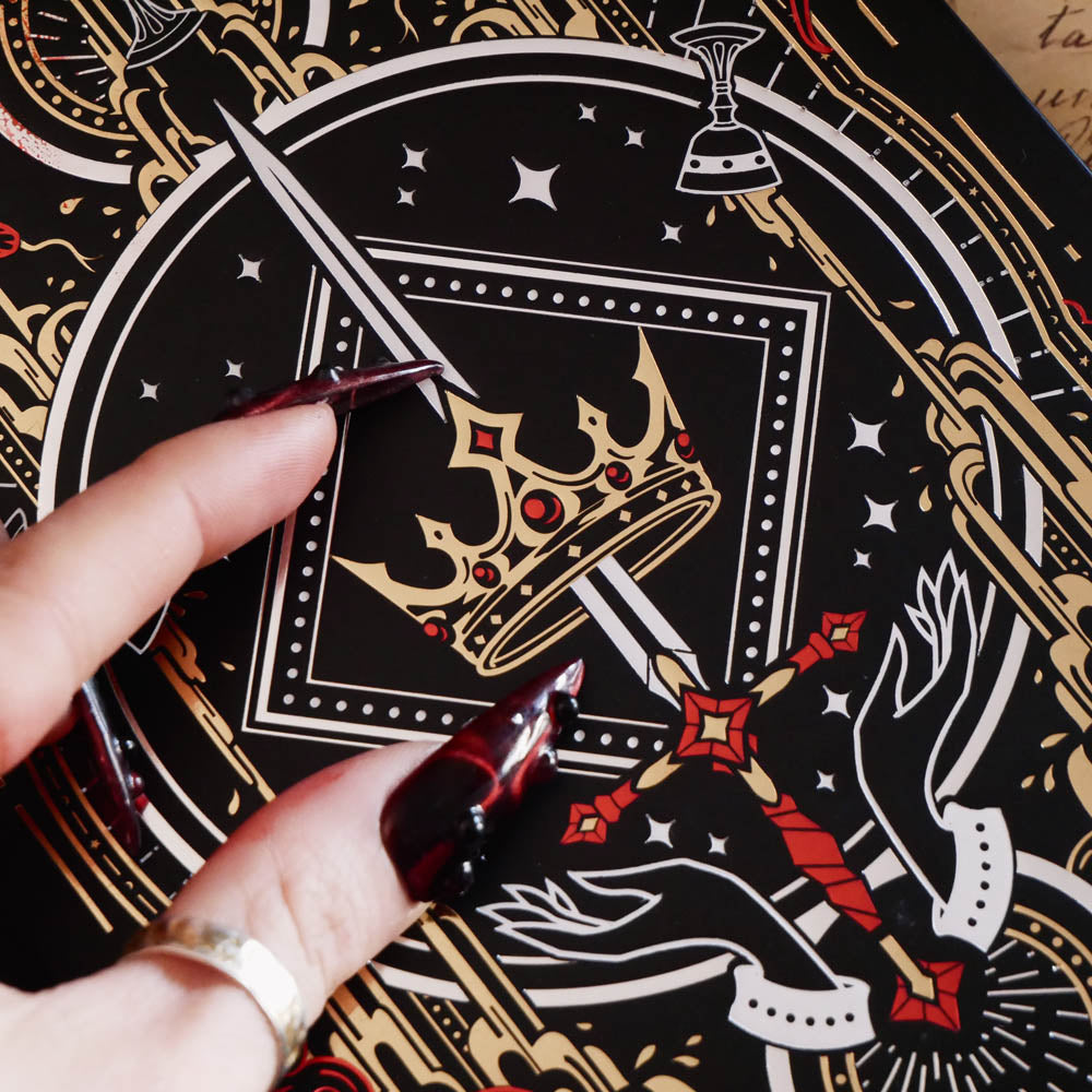 A detail of The Oath Breaker Journal sits on a red fabric background surrounded by handwritten notes, a candle and a crown. The black journal features intricate gold, red and white foiling with motifs inspired by fantasy books such as crowns, swords, goblets and snakes.