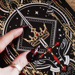 A detail of The Oath Breaker Journal sits on a red fabric background surrounded by handwritten notes, a candle and a crown. The black journal features intricate gold, red and white foiling with motifs inspired by fantasy books such as crowns, swords, goblets and snakes.