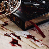 A detail of the Oath Breaker Journal sits surrounded by handwritten notes, a candle and a crown. The black journal features intricate gold, red and white foiling with motifs inspired by fantasy books such as crowns, swords, goblets and snakes. The gilded gold pages are shown with the red placeholder ribbon.