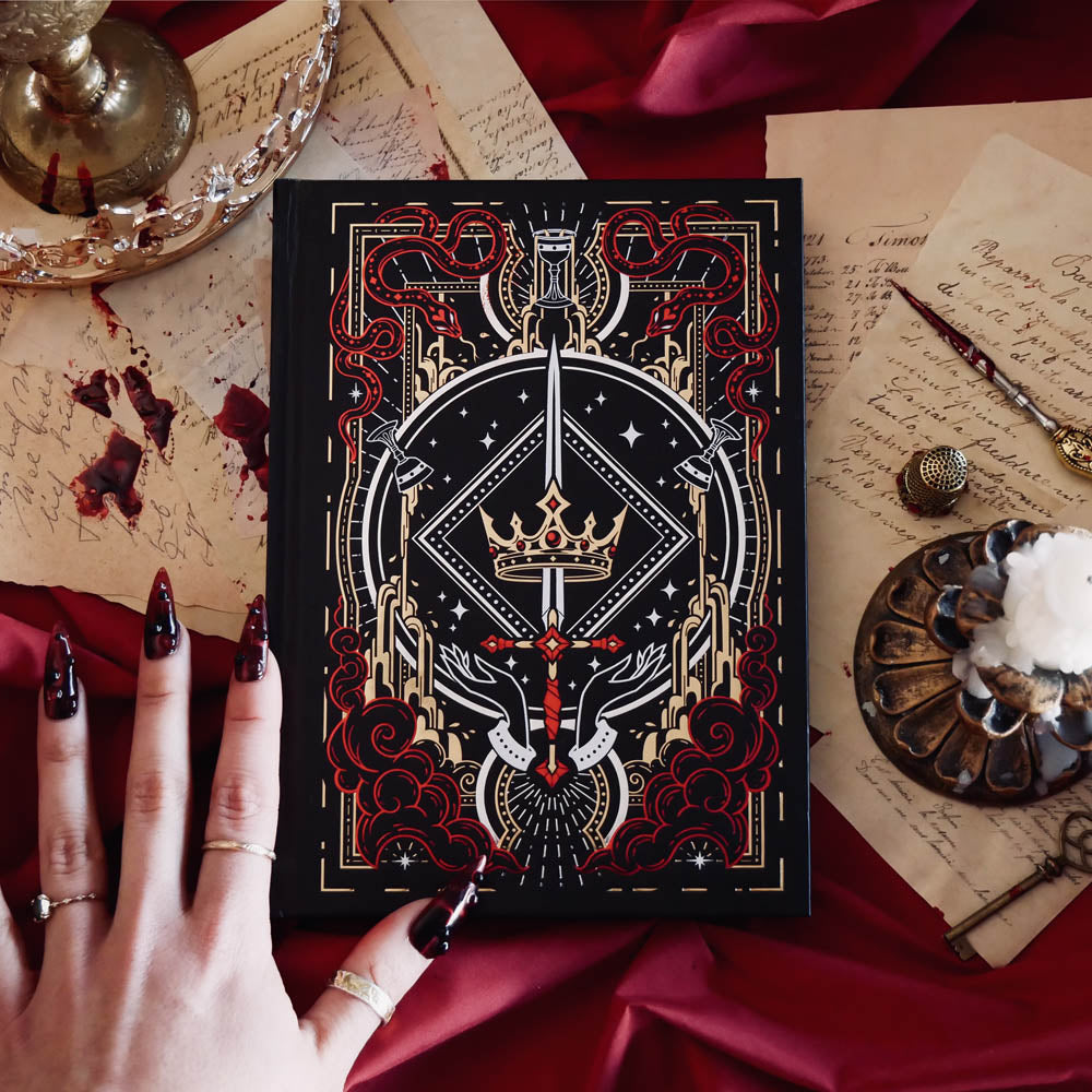 The Oath Breaker Journal sits on a red fabric background surrounded by handwritten notes, a candle and a crown. The black journal features intricate gold, red and white foiling with motifs inspired by fantasy books such as crowns, swords, goblets and snakes.